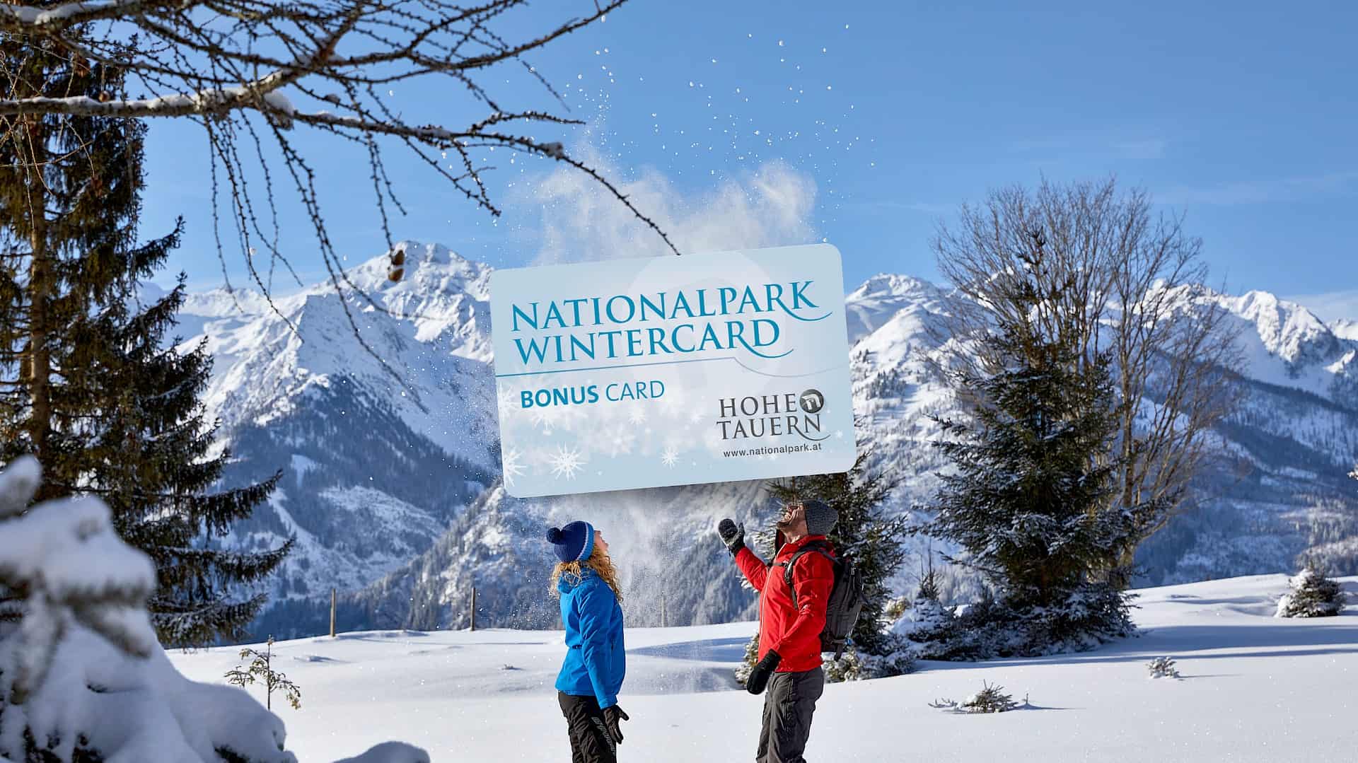 Zwei Winterwanderer im verschneiten Nationalpark Hohe Tauern freuen sich über die Nationalpark WinterCard Bonuskarte vor beeindruckender Alpenkulisse.