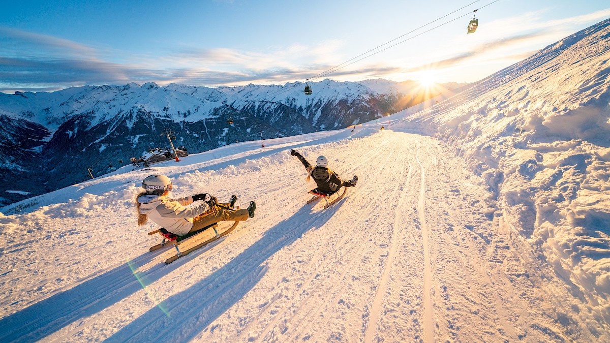 Twee mensen rodelen bij zonsondergang op de langste verlichte rodelbaan ter wereld – 14 km puur winterplezier. Ze dragen helmen en winterkleding en nemen lachend een bocht op het besneeuwde pad. Op de achtergrond de Alpen, met een gondellift erboven in het warme avondlicht.
