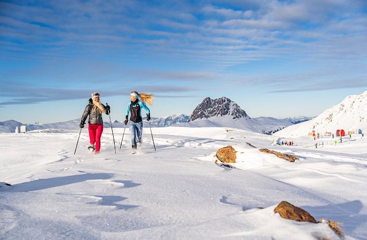 Zwei Frauen beim Schneeschuhwandern in der Wildkogel-Arena vor dem markanten Gipfel des Großen Rettensteins, bei Sonnenschein und glitzerndem Schnee.