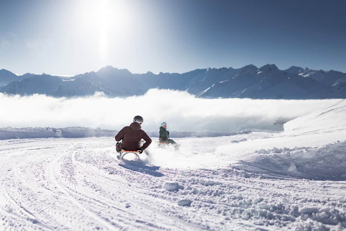 Two people sledding around a curve on the world’s longest floodlit toboggan run in the Wildkogel-Arena. The 14 km track winds through a stunning winter landscape with views of the snow-covered peaks of Hohe Tauern National Park, mist in the valley, and the sun shining above.