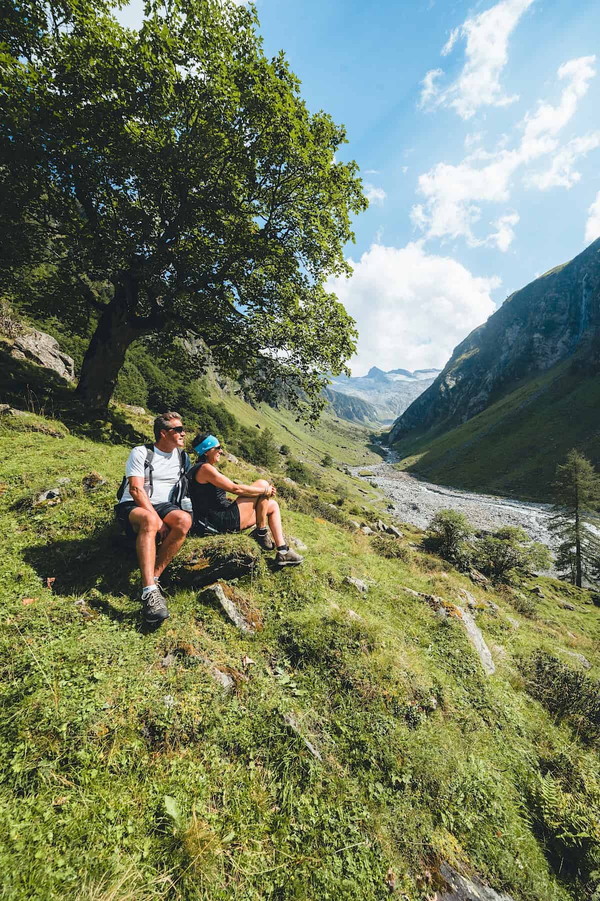 A couple enjoys a hiking break beneath a shady tree in Hohe Tauern National Park, overlooking a wild and romantic valley – active relaxation, nature experiences, and peace during a summer hike in the Wildkogel holiday region.