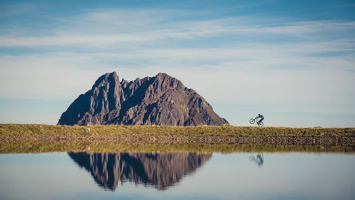 Ein Mountainbiker fährt entlang des Speichersees im Wander- und Bikeparadies Wildkogel, im Hintergrund spiegelt sich der markante Gipfel des Großen Rettensteins im klaren Wasser – perfektes Naturerlebnis für Aktivurlauber in den österreichischen Alpen.
